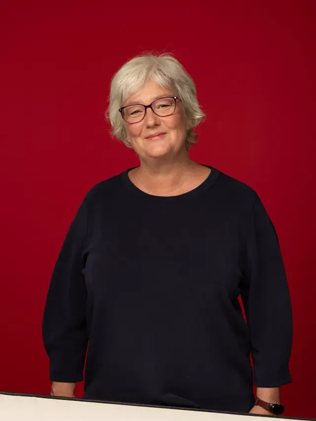 Inken Kuhn im dunkelblauen Pullover, lächelnd an einem Tisch stehend, vor rotem Hintergrund.